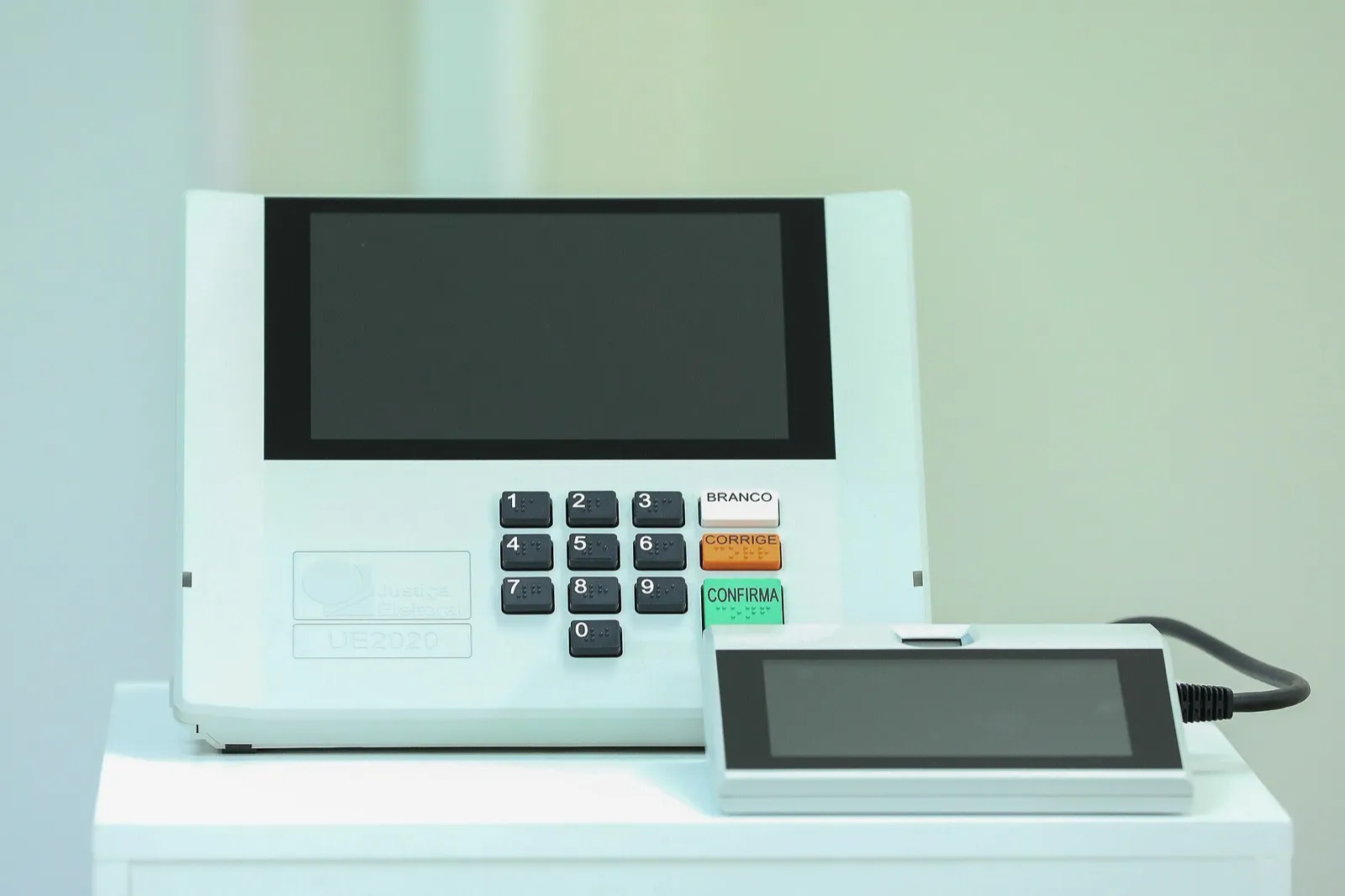 Urna eletrônica modelo UE2020, com tela apagada, teclado numérico e teclas BRANCO, CORRIGE e CONFIRMA. Ao lado, um visor menor ligado por cabo.
