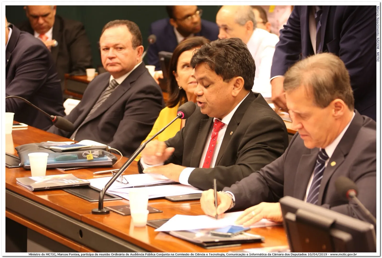 Parlamentares sentados à mesa de uma comissão da Câmara durante reunião com microfones e documentos.