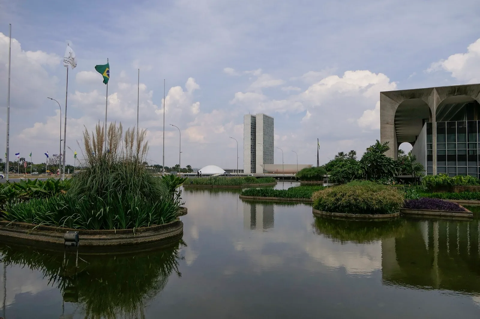 Espelho d'água com gramados em primeiro plano e, ao fundo, as duas torres do Congresso Nacional e uma cúpula branca, em Brasília, sob céu com nuvens.