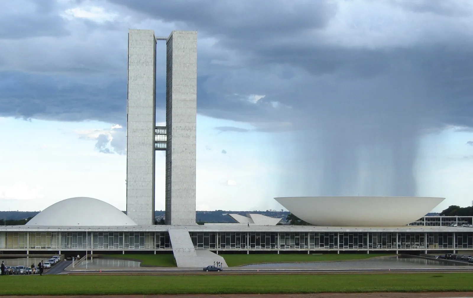 Sede do Congresso Nacional em Brasília, com as duas torres no centro, a cúpula do Senado à esquerda e a da Câmara à direita, sob céu com chuva ao fundo.