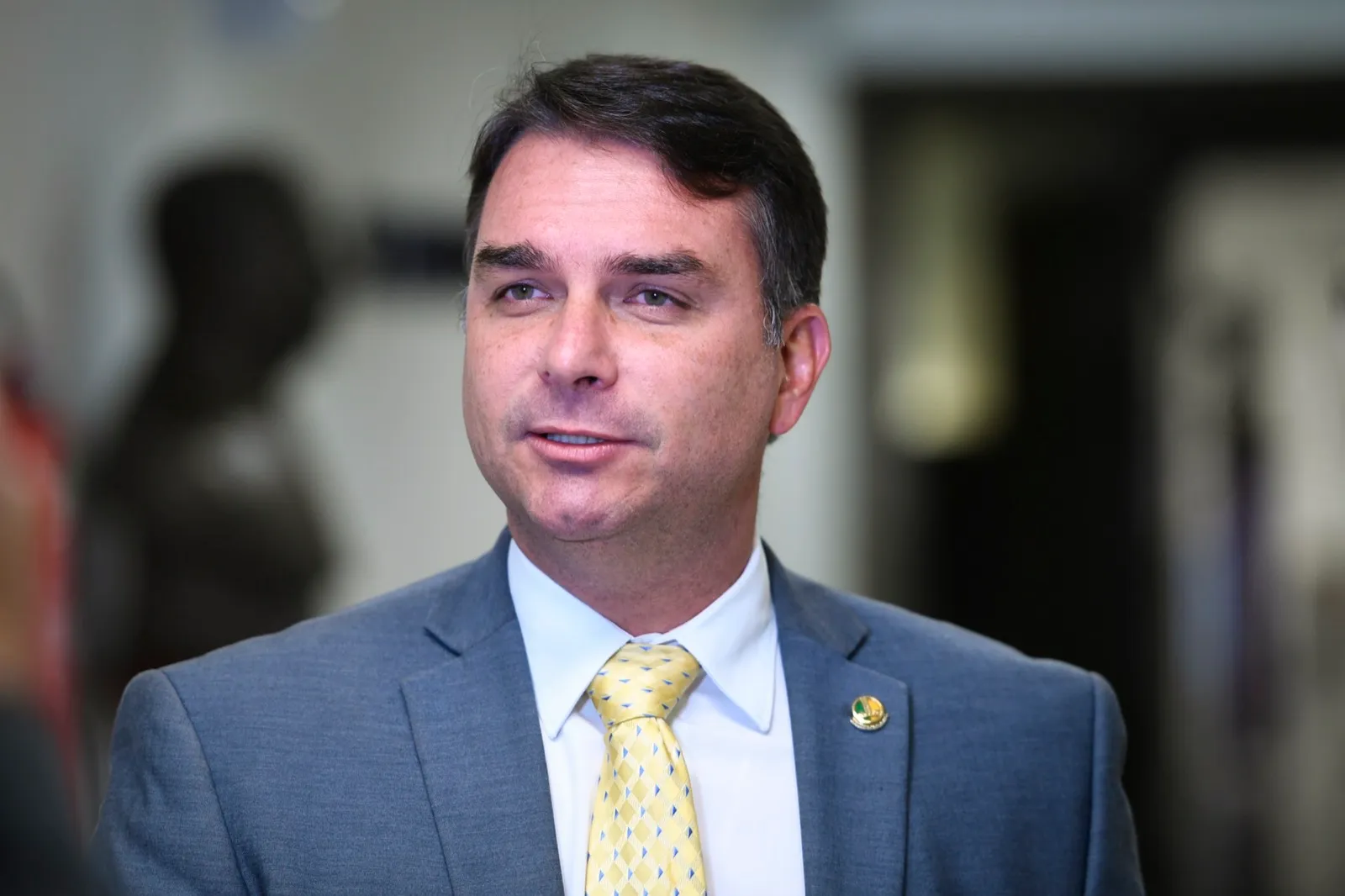 Flávio Bolsonaro, de terno cinza e gravata amarela, em primeiro plano durante entrevista num corredor do Senado.