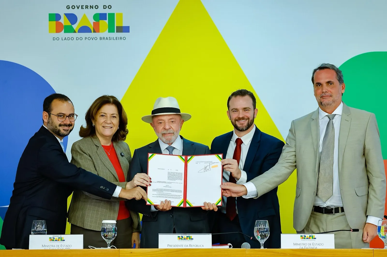 Lula segura, no Palácio do Planalto, a medida provisória do Novo Desenrola Brasil, ao lado de ministros, entre eles o ministro da Fazenda, que aparece de terno claro à direita.