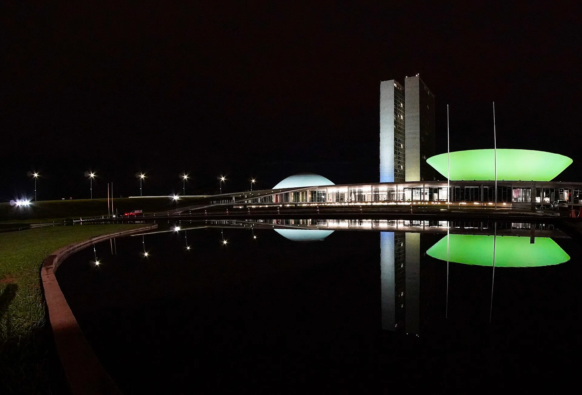 O Congresso Nacional à noite, com as duas torres, a cúpula branca e a cúpula invertida iluminada de verde, refletidas no espelho d'água.