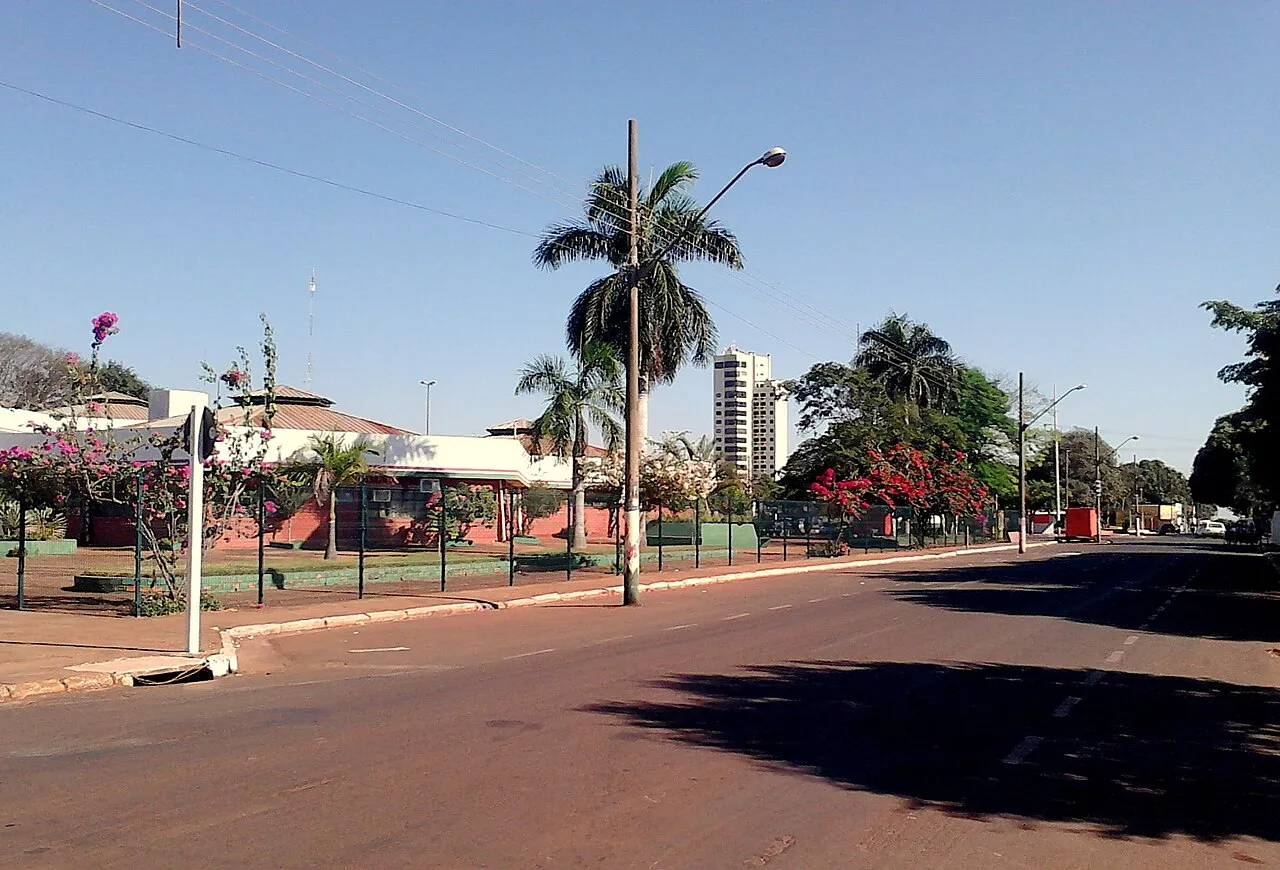 Rua larga e vazia em Tangará da Serra, com palmeiras e árvores floridas, um prédio térreo de telhado vermelho à esquerda e um edifício alto ao fundo, sob céu azul.