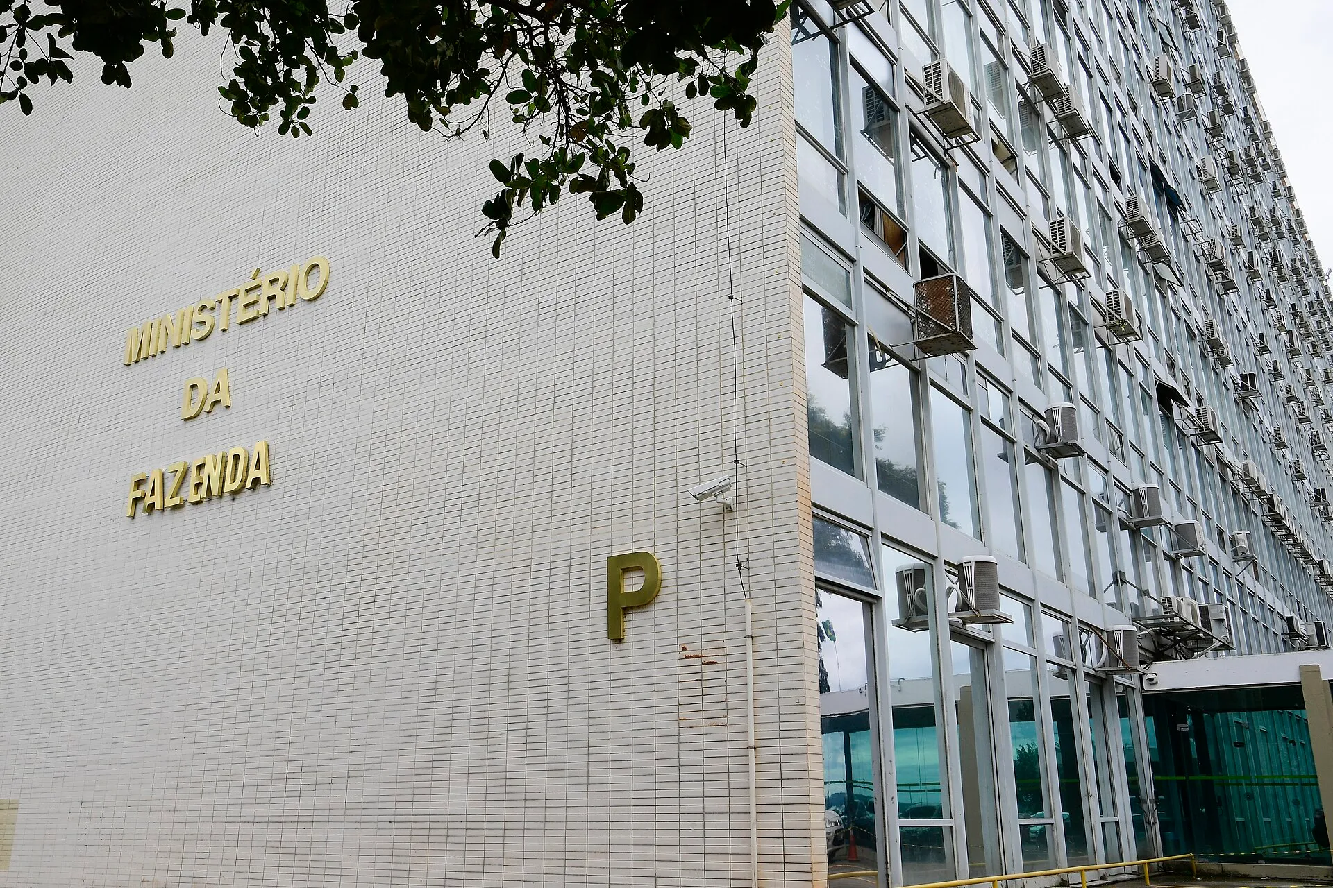 Fachada do bloco P do Ministério da Fazenda, em Brasília, com paredes de azulejos brancos, letras douradas e janelas de vidro, sob céu nublado.