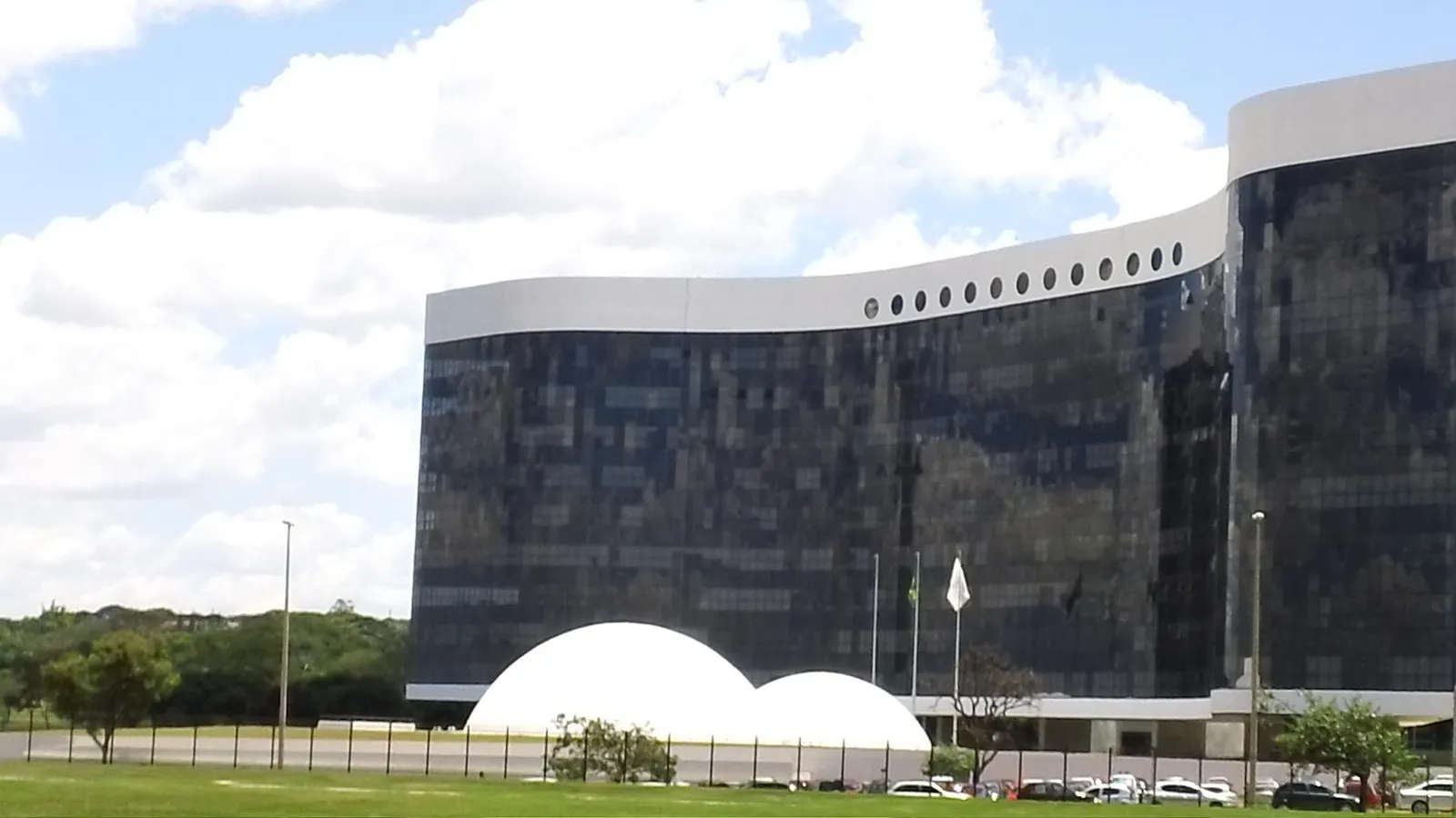 Fachada espelhada e curva do edifício-sede do Tribunal Superior Eleitoral, em Brasília, com duas cúpulas brancas em primeiro plano e céu com nuvens.