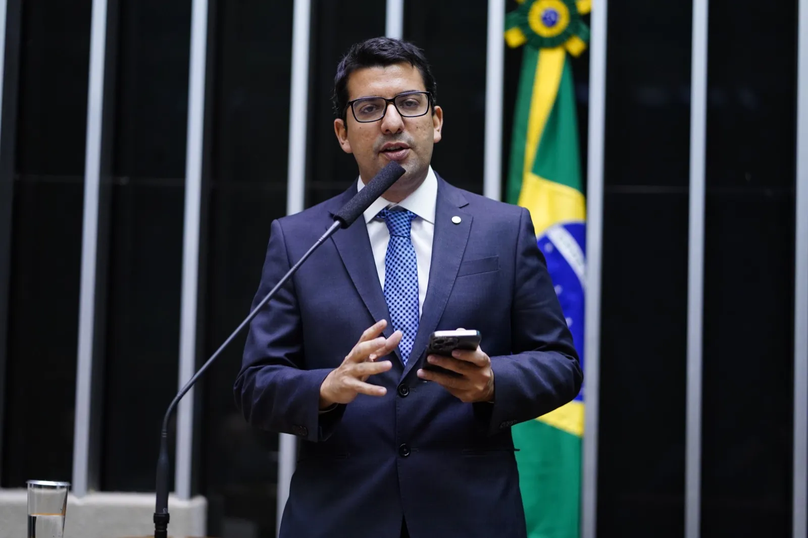 O deputado Marcelo Queiroz fala ao microfone no plenário da Câmara, com celular em uma das mãos.