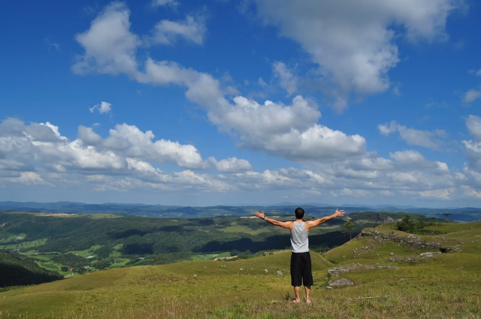 Pessoa de costas observa um vale com morros e vegetação em Alfredo Wagner, Santa Catarina, sob céu azul com nuvens.