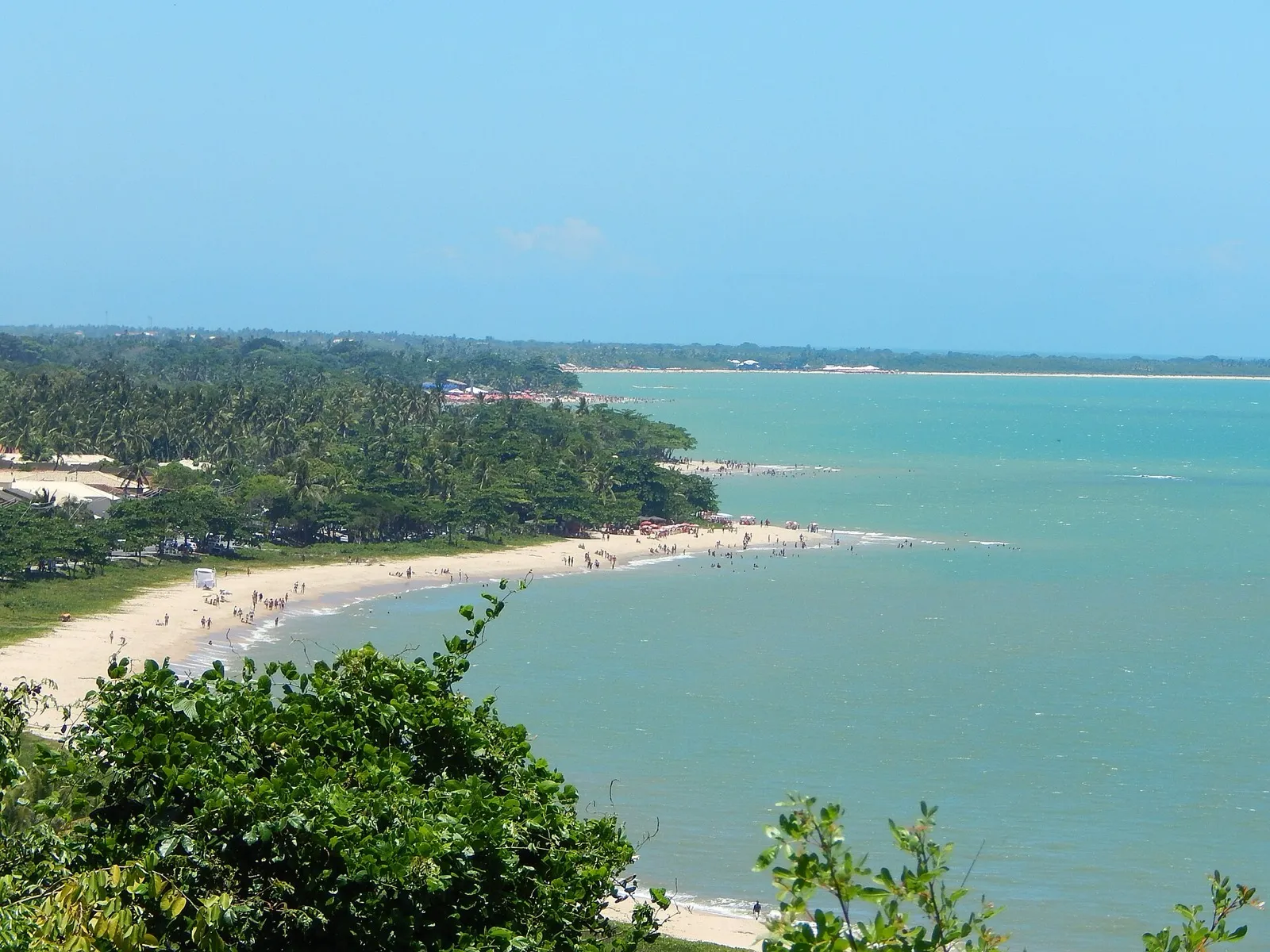Vista do alto de uma praia de areia clara e mar azul-esverdeado em Porto Seguro, na Bahia, com vegetação e banhistas.