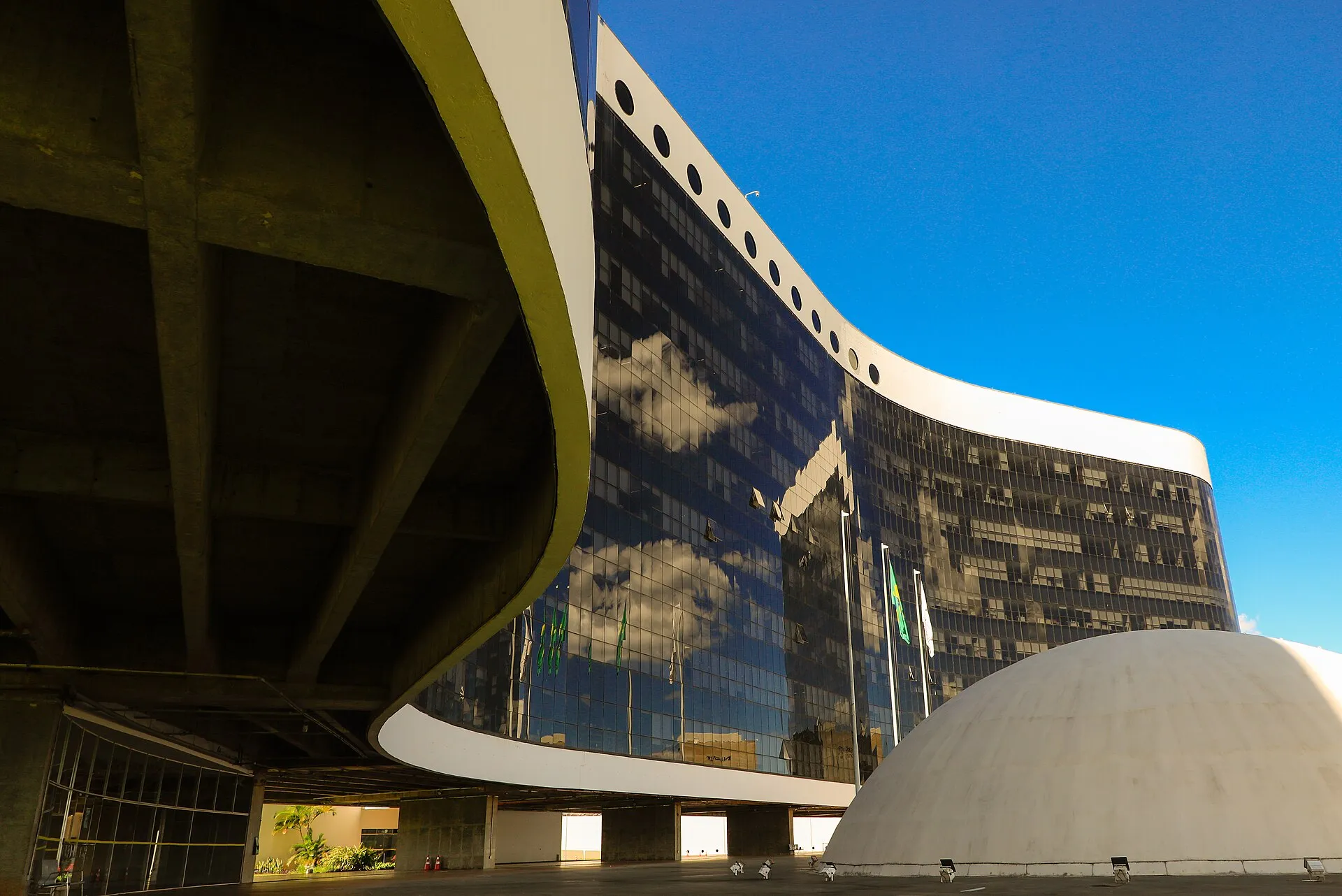 Fachada curva de vidro escuro da sede do Tribunal Superior Eleitoral, em Brasília, com a cúpula branca do projeto de Oscar Niemeyer em primeiro plano, sob céu azul.