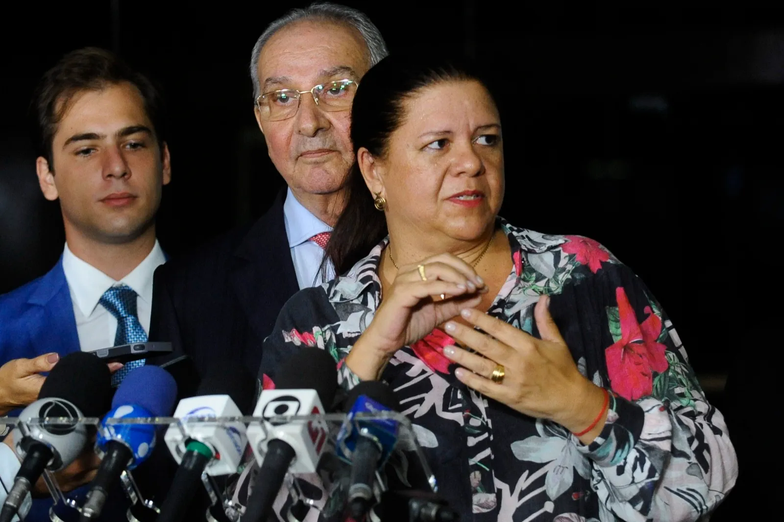 A deputada Laura Carneiro fala diante de microfones durante entrevista no Senado Federal, com dois homens atrás dela.