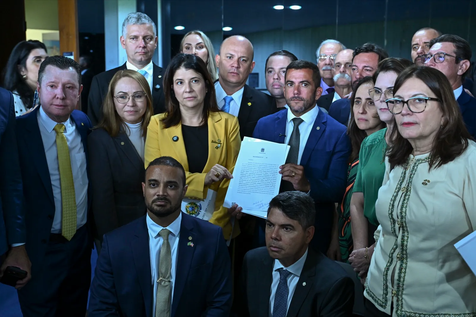 Grupo de deputados posa no Congresso; ao centro, uma deputada de blazer amarelo e um deputado de terno azul seguram um requerimento.
