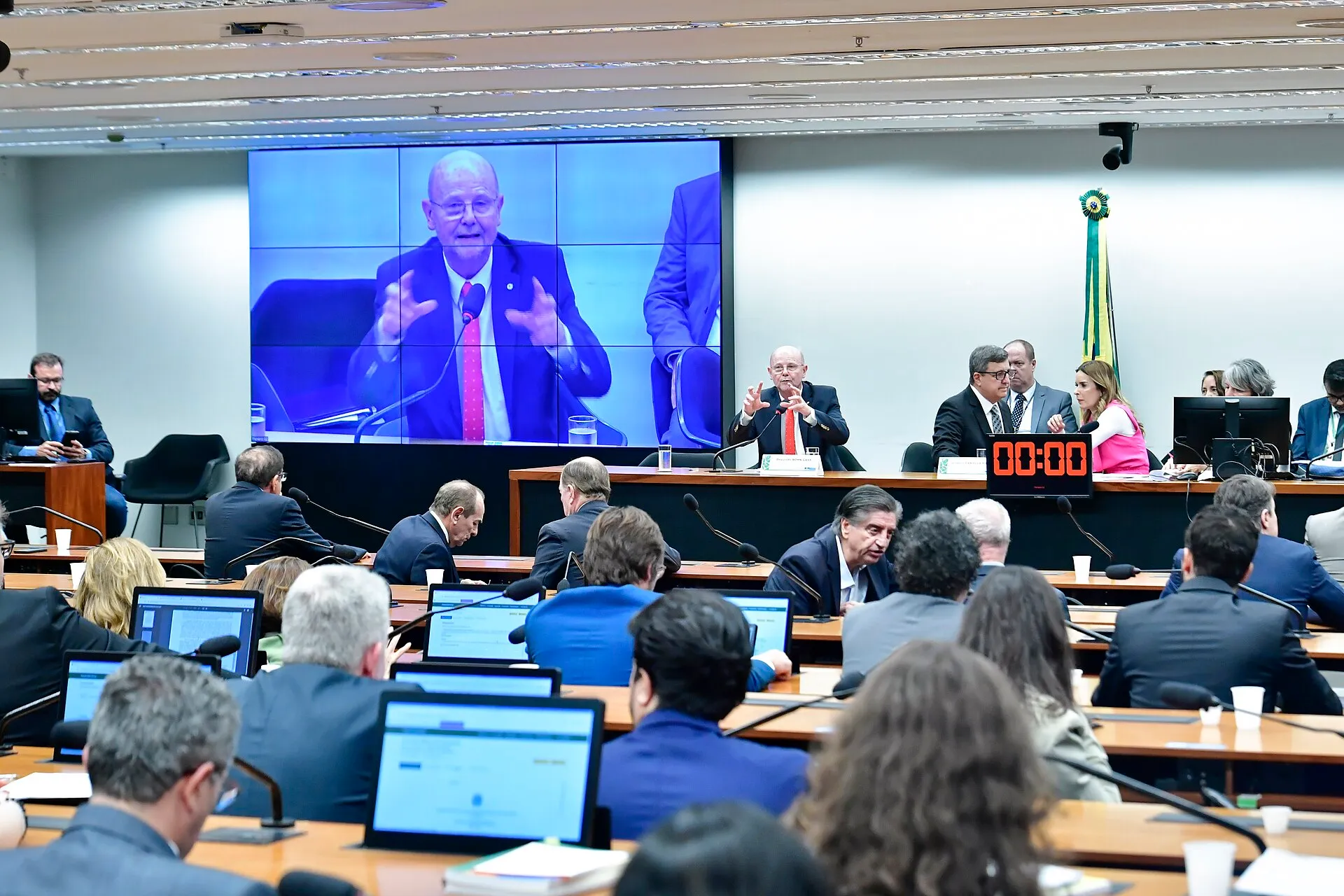 Sala de reunião da Comissão Mista de Orçamento, com parlamentares sentados diante de notebooks e, ao fundo, a mesa diretora e um telão que mostra um orador.