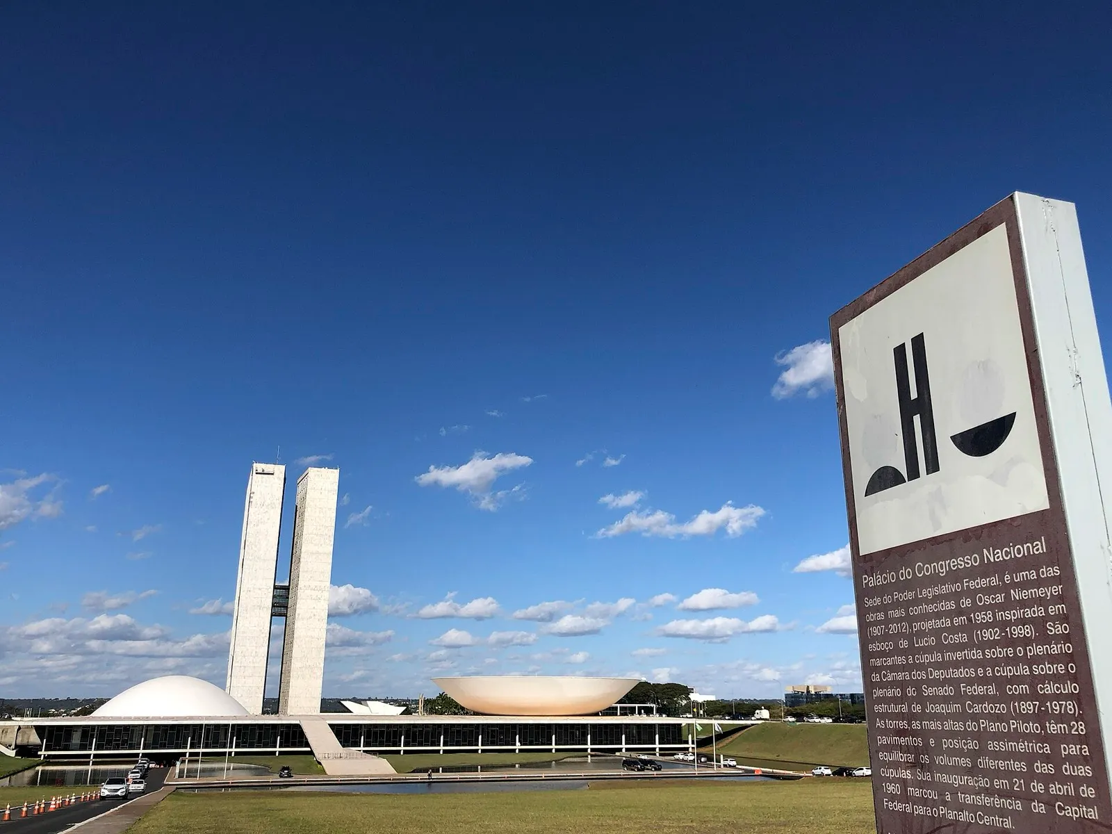 Palácio do Congresso Nacional, em Brasília, com as duas torres e as cúpulas do Senado e da Câmara ao fundo, sob céu azul, e uma placa informativa em primeiro plano.