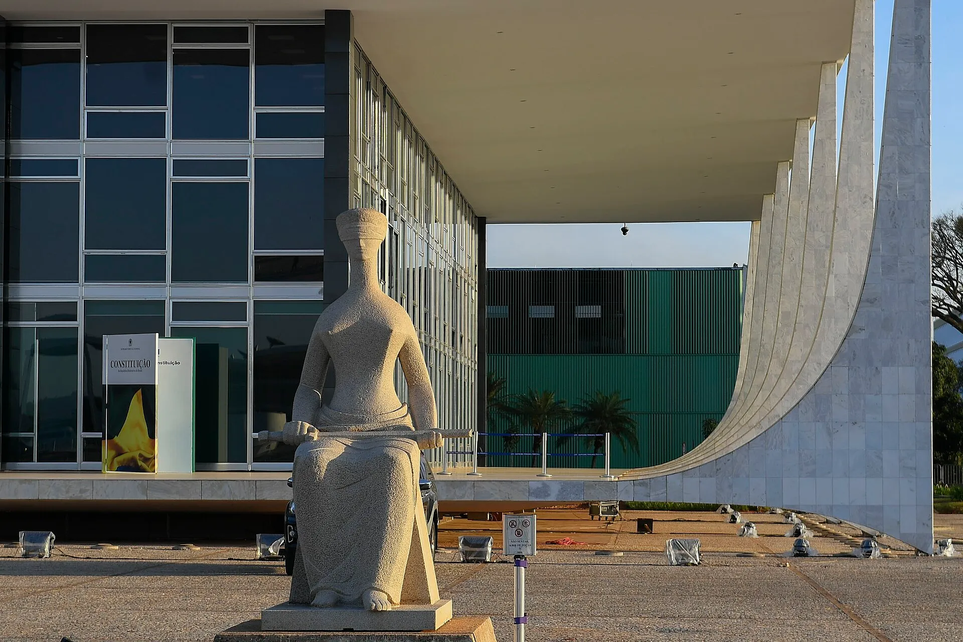 Estátua da Justiça, de olhos vendados e com uma espada no colo, em frente à fachada de vidro e às colunas curvas de mármore do Supremo Tribunal Federal, em Brasília.