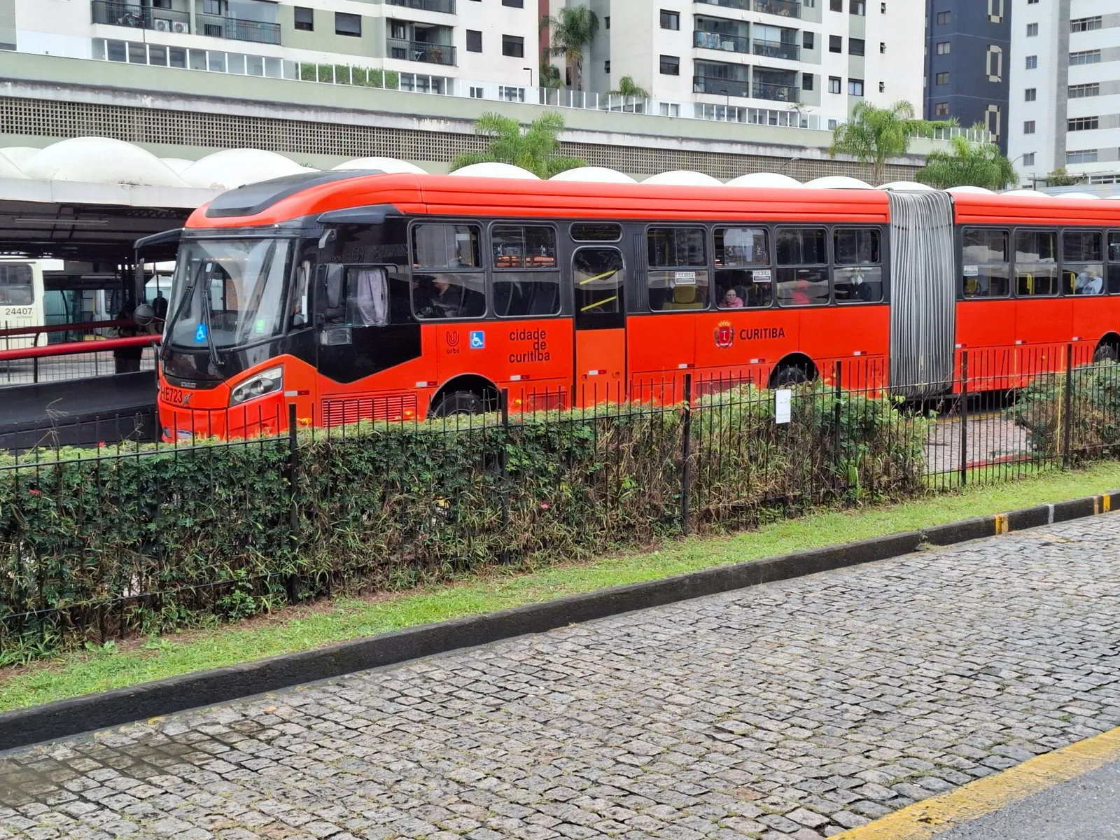 Ônibus biarticulado laranja com as inscrições Cidade de Curitiba e Urbs parado no Terminal Cabral, atrás de uma cerca viva.