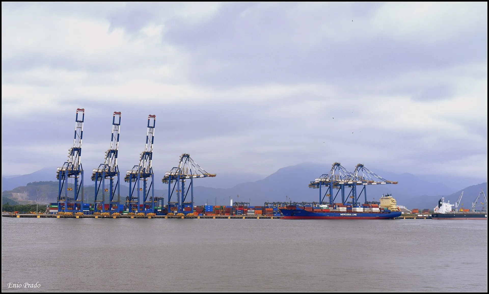 Guindastes azuis de um terminal de contêineres do Porto de Santos, com um navio atracado e a água do canal em primeiro plano.