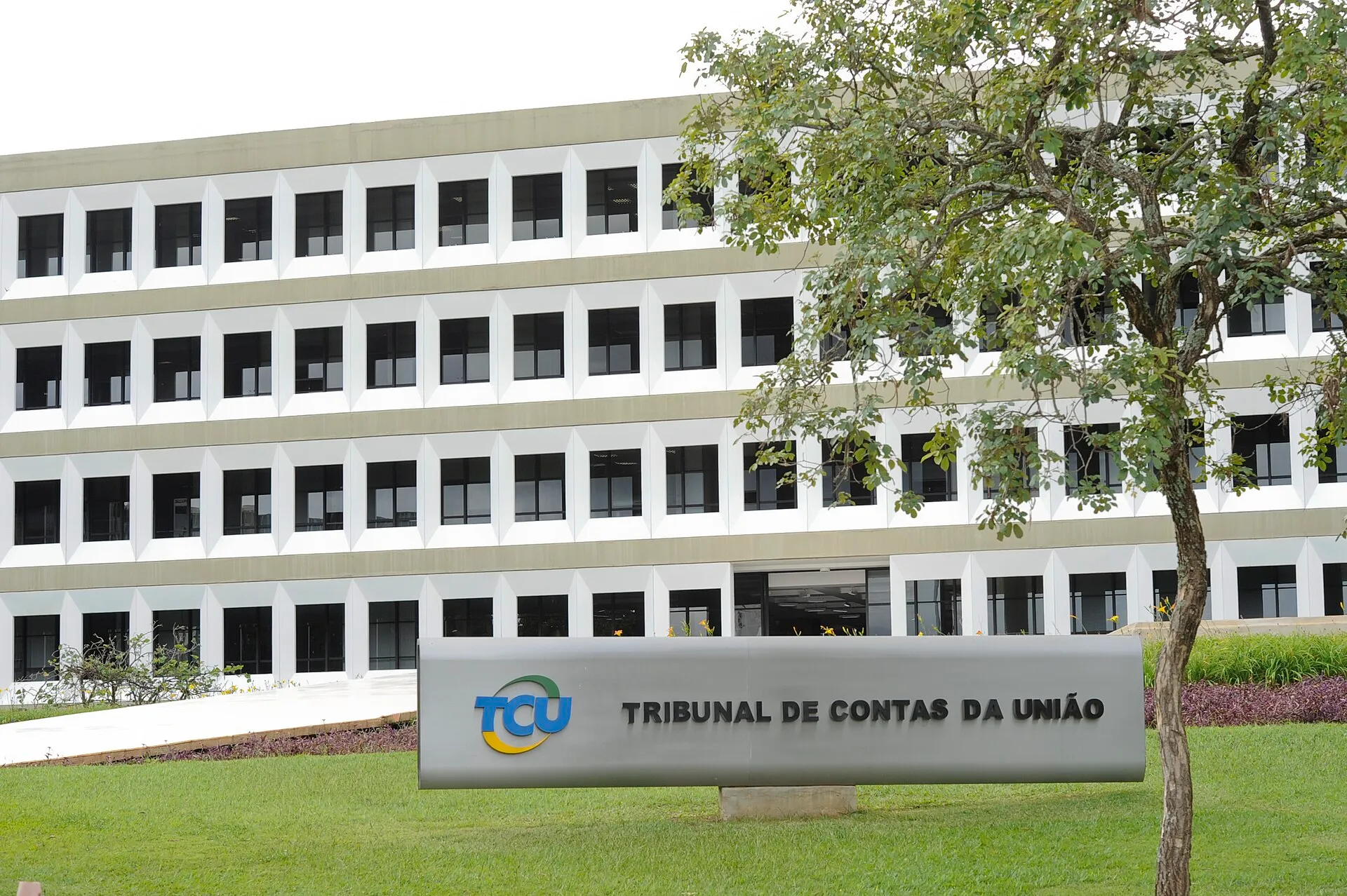 Fachada branca de vários andares do edifício-sede do Tribunal de Contas da União, em Brasília, com a placa do órgão no gramado e uma árvore à direita.