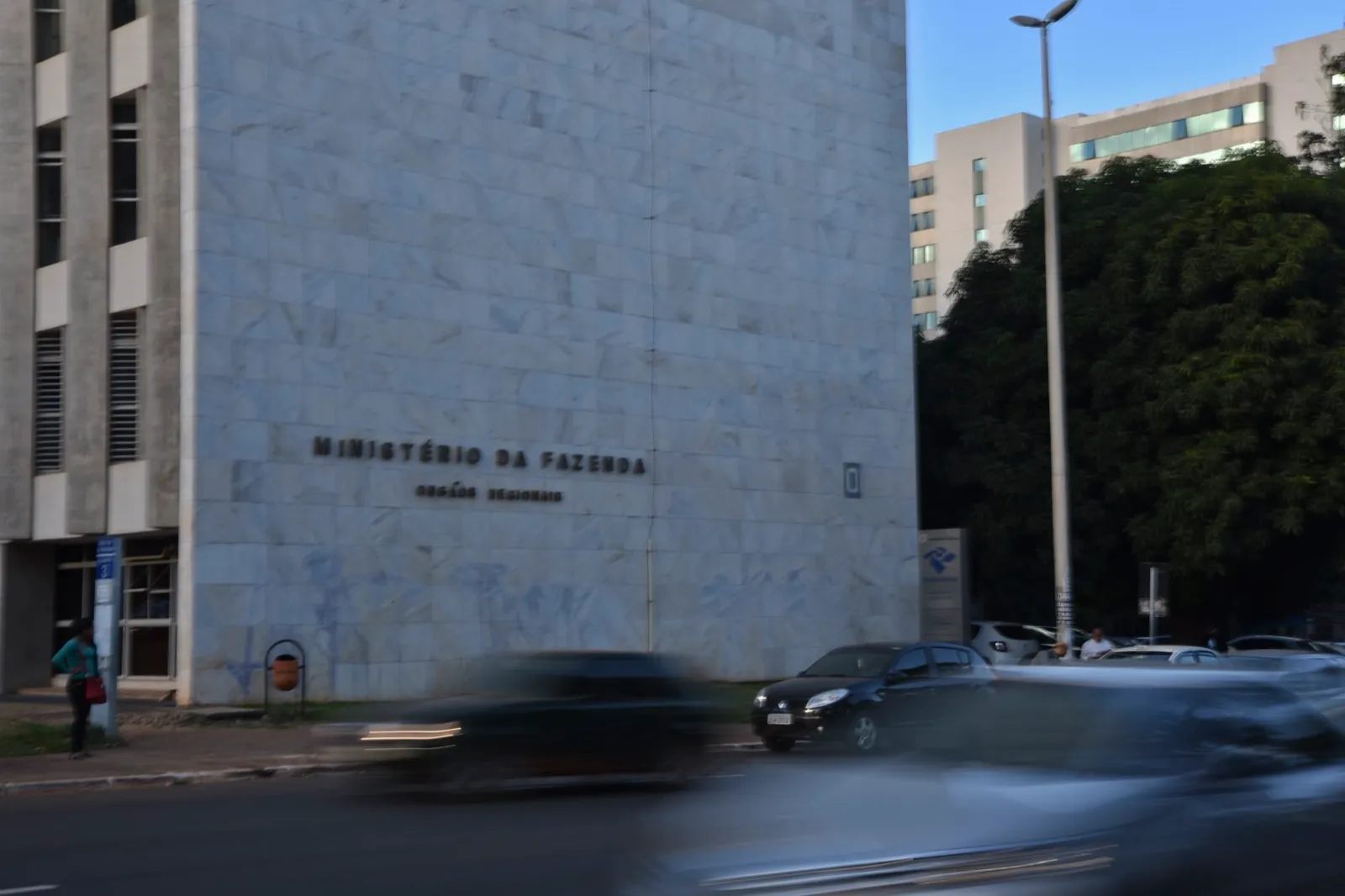 Fachada de concreto do Ministério da Fazenda, em Brasília, com o nome do órgão e veículos passando em primeiro plano.